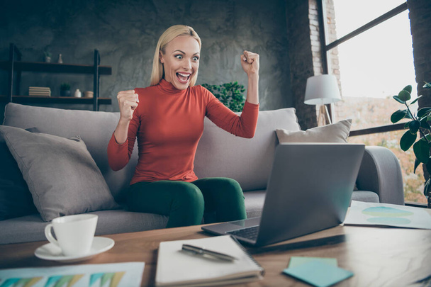 Photo of crazy blond business lady raising fists up celebrating first investment startup project freelancer wear casual orange pullover sitting sofa table notebook indoors - Foto, Imagem
