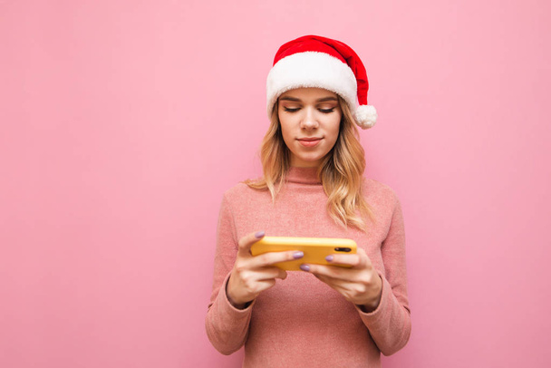 Portrait of a cute girl in a Christmas hat and a pink sweater isolated on a pink background, uses a smartphone, looks at the screen with a serious face, Calm girl plays mobile games on a smartphone - Photo, Image