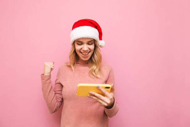Retrato de chica feliz en sombrero de Navidad jugando juegos móviles en el teléfono inteligente, regocijo alegre para ganar en el fondo rosa, mira a la pantalla del teléfono inteligente y sonríe. Concepto de Navidad y Año Nuevo
. - Foto, imagen