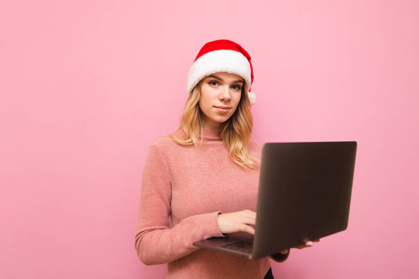 Girl worker in a pink sweater and Christmas hat stands with a laptop in hand on a pink background and looks into the camera with a serious face.Lady in Santa hat with laptop in hand,isolated - Photo, image