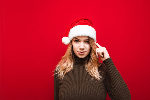 Atraktivní dívka v Santa klobouku stojí na červeném pozadí, dívá se do kamery a ukazuje gesto přemýšlet, dotýká se prstem spánku. Přemýšlej o tom.. - Fotografie, Obrázek