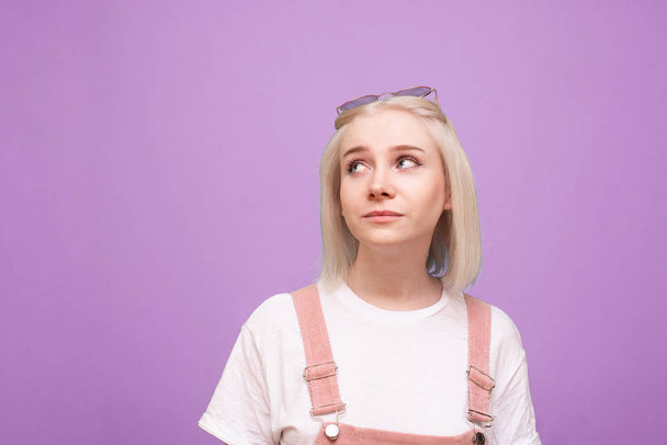 Cute girl in a bright casual dress, wearing a white T-shirt, pink sunglasses and dress, looks out to the empty place with a funny face. Positive teen girl on a purple background looks to the side - Photo, Image