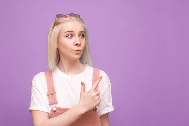 Portrait of surprised sweet lady showing thumb to the side, isolated on a purple background. Emotional girl looks away with a shocking face and shows her finger in a place for advertising. - Photo, Image