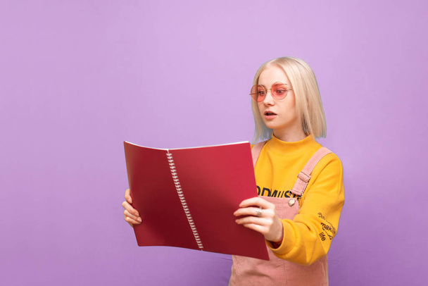 Cute girl blonde in pink glasses and bright casual clothes, looking face to face with a serious note on a purple background. Portrait of a funny student with a notebook in his hands, reads. - Photo, Image