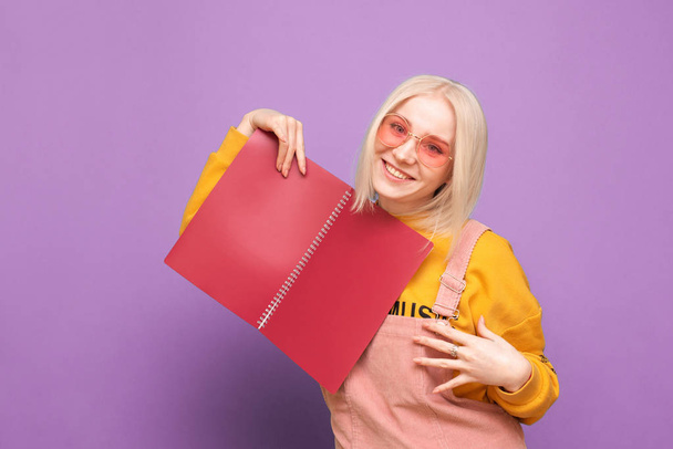 Happy girl blonde in bright clothes and pink glasses poses with a broom in her hands on a purple background, looks at the camera and smiles. Portrait of a funny teen with a notebook in his hands. - Photo, Image