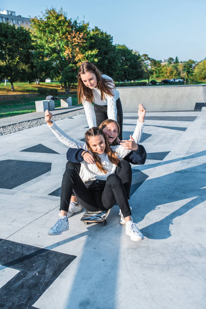 Three teenage girls schoolgirls ride a skateboard after school, happy smiling, emotions of joy, delight and fun, warm sweaters in autumn on street in city. Best friends girlfriends teens. - Photo, Image