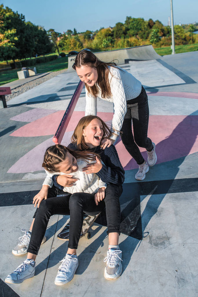 Three teenage girls schoolgirls ride skateboard after school, happy smiling, emotions of joy, delight and fun, warm sweaters in autumn on street city. Best friends girlfriends teens. - Фото, изображение