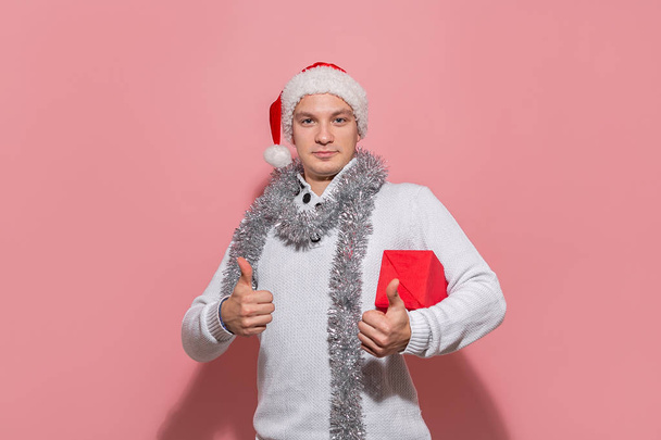 Man in a white sweater and red Santa hat holding Christmas gift in a red box showing thumb up isolated on pink background. - Photo, image