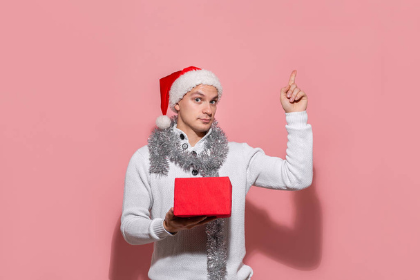 Hombre con un suéter blanco y un sombrero rojo de Santa señalando con el dedo el espacio de copia hacia arriba
. - Foto, imagen