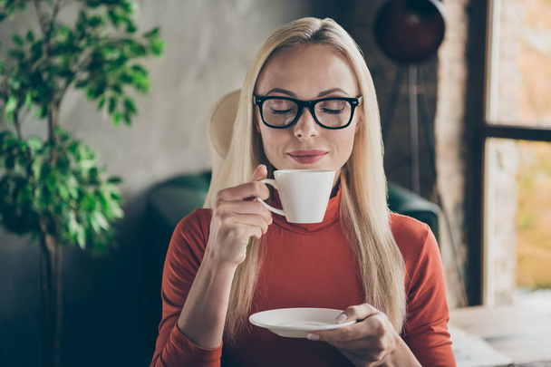 Close up photo of peaceful worker woman in orange turtleneck have rest relax hold white coffee cup smell aroma enjoy in loft office workstation red turtlneck - Foto, Imagem