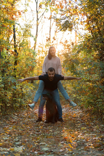 Young beautiful couple and their labrador in the Park. A man holds a woman on his back, and they spread their arms. Rays of setting sun break through the branches of trees. - Foto, afbeelding