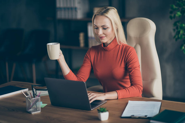 Photo of blonde haired cheerful positive programmer working on her new software for corporation she works for holding mug sitting at desktop - Fotó, kép