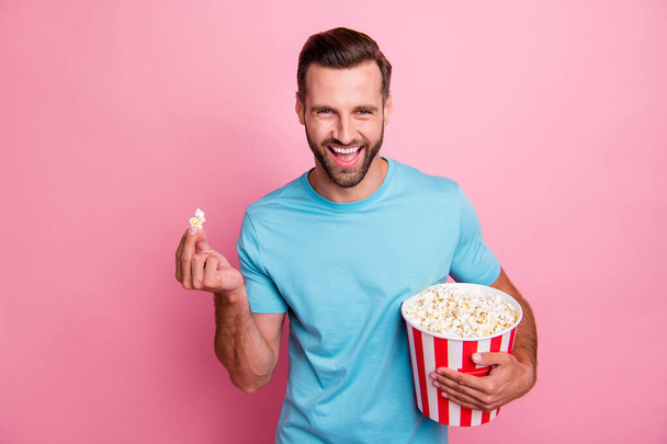 Photo of cheerful positive man watching tv series holding bucket of popcorn tasting smiling toothily comedy isolated in blue t-shirt over pastel color background - Fotografie, Obrázek