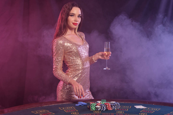 Woman in golden dress holding chip and champagne while posing at playing table in casino. Black, smoke background. Gambling, poker. Close-up. - Photo, image
