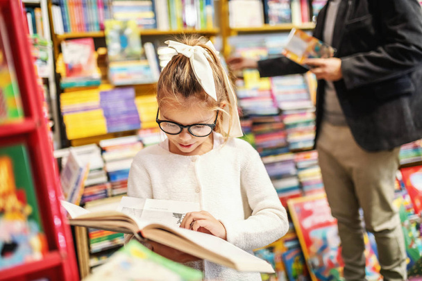 Adorable curious caucasian little girl with eyeglasses standing in bookstore and reading interesting book. All around are books on shelves. - Φωτογραφία, εικόνα