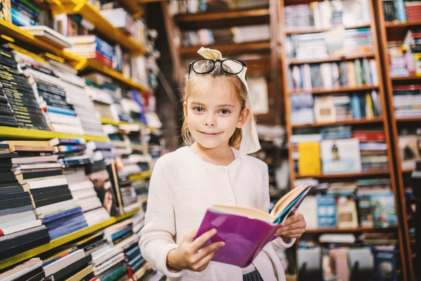 Adorable curious caucasian little girl with eyeglasses standing in bookstore and holding interesting book while looking at camera. All around are books on shelves. - Foto, imagen