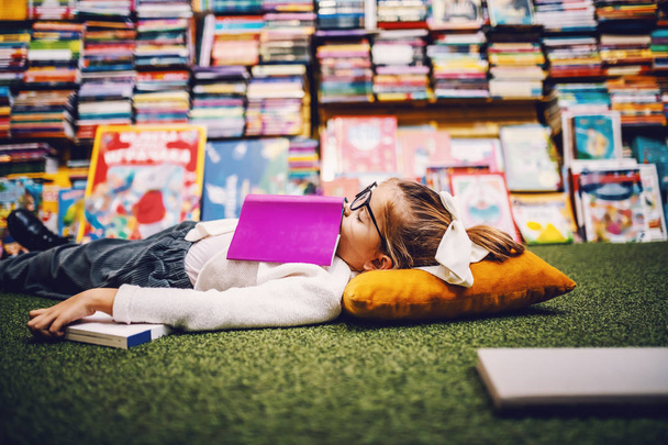Encantadora niña rubia caucásica con cola de caballo tirada en el suelo en la librería con libro en el pecho y durmiendo. Todo alrededor hay libros para niños.
. - Foto, imagen