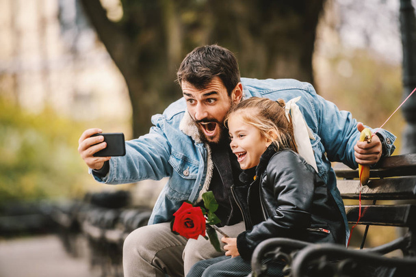 Felice sorridente padre caucasico barbuto seduto sulla panchina nel parco con la sua amata figlia e prendendo selfie. Ora di autunno
. - Foto, immagini