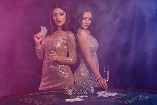 カジノのテーブルでポーズをとりながらカードを見せる2人の豪華な女性。カラフルなバックライトで黒、煙の背景。ギャンブル、ポーカー。閉鎖. - 写真・画像