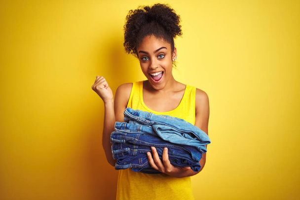 Young african american woman holding stack of jeans over isolated yellow background screaming proud and celebrating victory and success very excited, cheering emotion - Valokuva, kuva