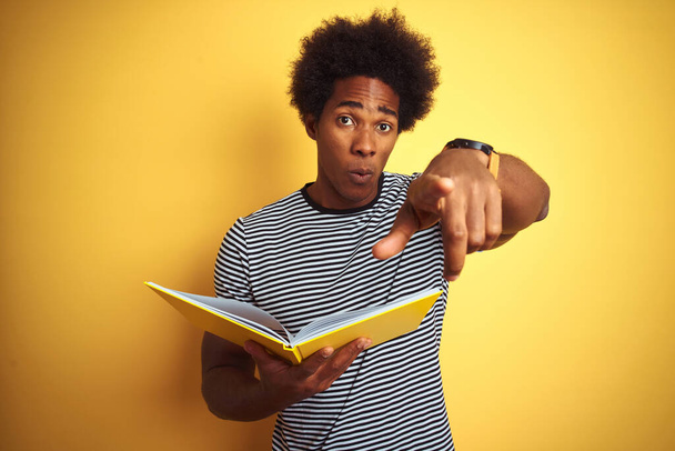 Афро-американский студент читает книгу стоя на изолированном желтом фоне указывая пальцем на камеру и на вас, знак руки, позитивный и уверенный жест спереди
 - Фото, изображение