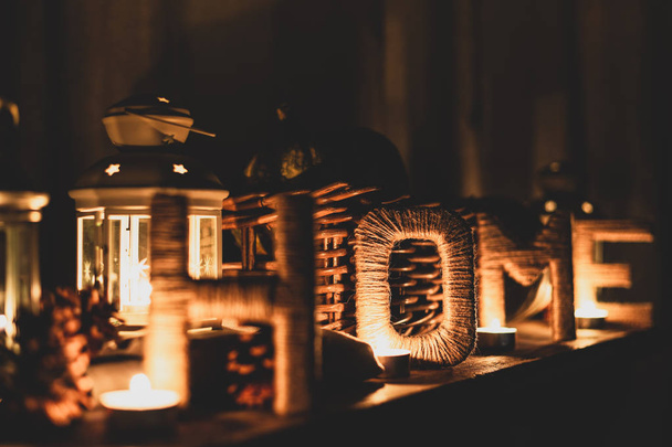 Decorative composition with inscription HOME, pine cones, candles and lanterns. Letters covered with twine in candlelight. Selective focus on letter O. - Valokuva, kuva