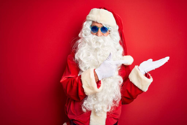 Середній вік чоловіка, одягненого в костюм Санта Клауса і сонячні окуляри над ізольованим червоним фоном, вражений і посміхаючись камери, представляючи з рукою і вказуючи пальцем.. - Фото, зображення