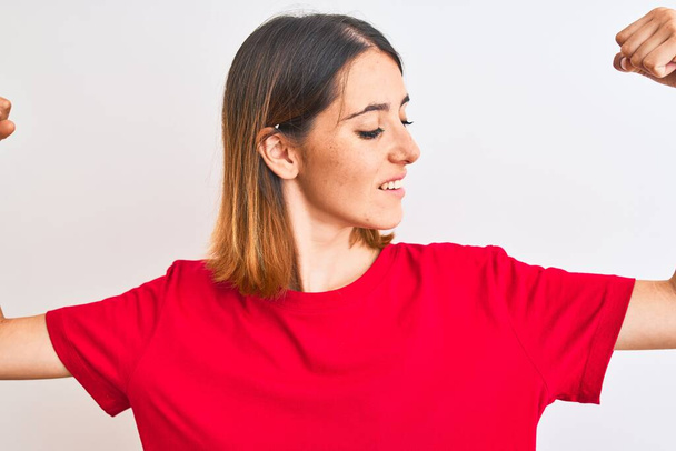 Όμορφη κοκκινομάλλα γυναίκα φορώντας casual κόκκινο t-shirt πάνω από απομονωμένη φόντο δείχνει τους μυς των χεριών χαμογελώντας υπερήφανοι. Έννοια καταλληλότητας. - Φωτογραφία, εικόνα
