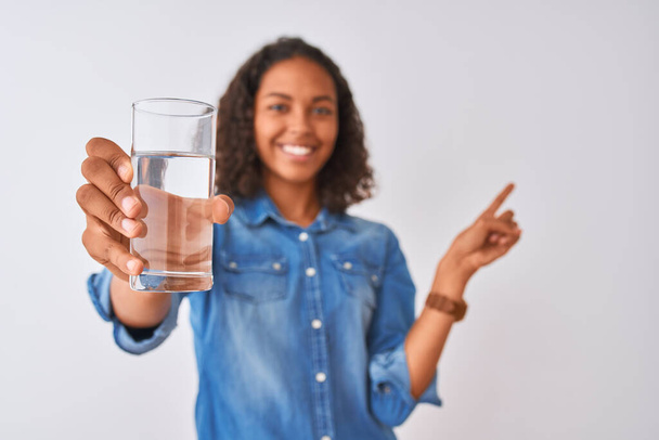 Mladá brazilská žena drží sklenici vody stojící nad izolovaným bílým pozadím velmi šťastný ukazující rukou a prstem na stranu - Fotografie, Obrázek
