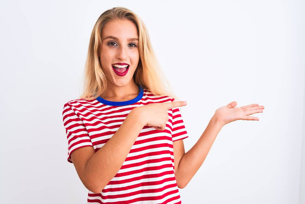 Молодая красивая женщина в красной полосатой футболке, стоящая на изолированном белом фоне удивлена и улыбается в камеру, представляя с рукой и указывая пальцем
. - Фото, изображение