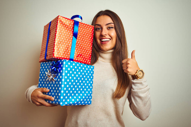 Young beautiful woman holding birthday gifts standing over isolated white background happy with big smile doing ok sign, thumb up with fingers, excellent sign - Photo, Image