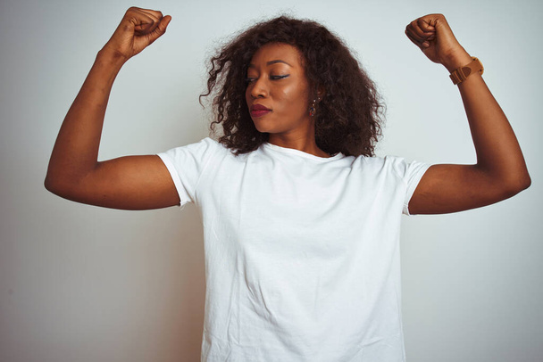 Genç Afro-Amerikalı kadın tişört giyip izole edilmiş beyaz arka planda duruyor ve gururla gülümseyen kol kaslarını gösteriyor. Fitness konsepti. - Fotoğraf, Görsel