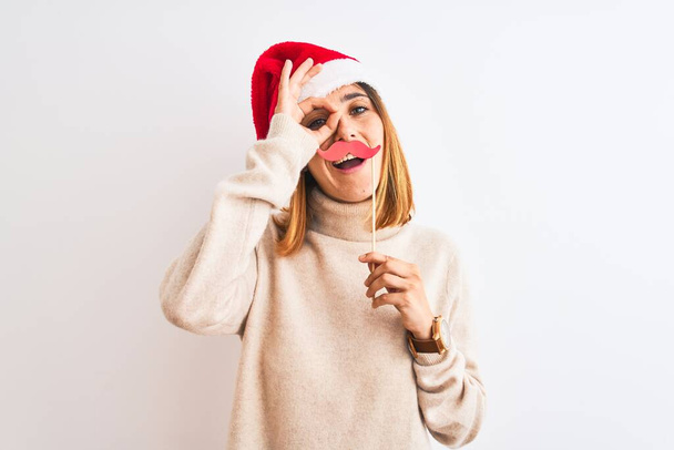 Gyönyörű vörös hajú nő visel karácsonyi kalap visel színlelt bajusz felett elszigetelt háttér boldog arc mosolyog csinál rendben jel kéz a szemen keresztül néz az ujjakon - Fotó, kép