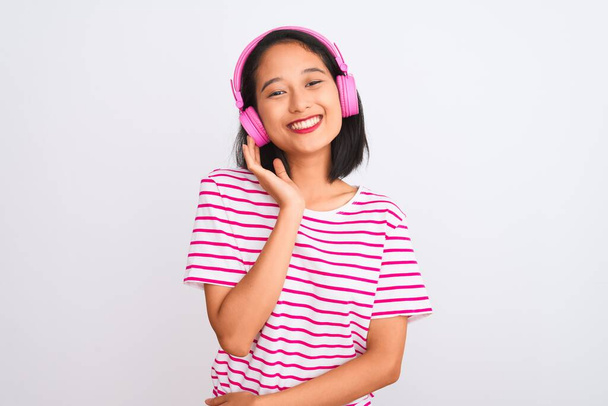 Красивая китаянка слушает музыку с помощью наушников на изолированном белом фоне с счастливым лицом стоя и улыбаясь с уверенной улыбкой показывая зубы
 - Фото, изображение