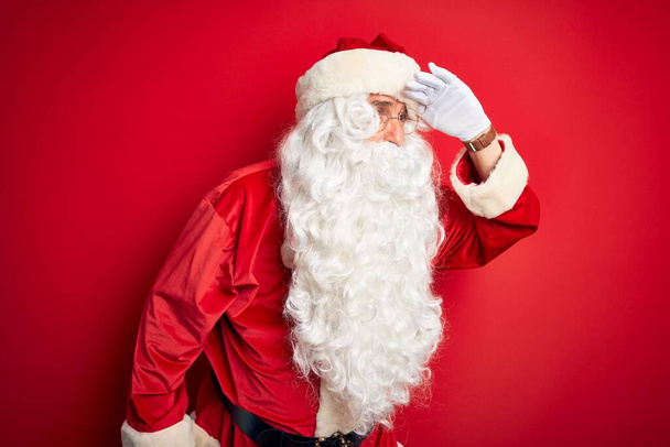 Middle age handsome man wearing Santa costume standing over isolated red background very happy and smiling looking far away with hand over head. Searching concept. - Photo, Image