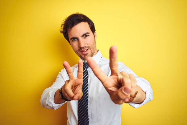Jonge knappe zakenman draagt elegant shirt en stropdas over geïsoleerde gele achtergrond glimlachend naar de camera kijkend met vingers die overwinningstekens doen. Nummer twee. - Foto, afbeelding