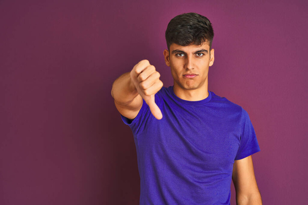 Hombre indio joven con camiseta de pie sobre un fondo púrpura aislado que parece infeliz y enojado mostrando rechazo y negativo con el gesto de los pulgares hacia abajo. Mala expresión
. - Foto, Imagen