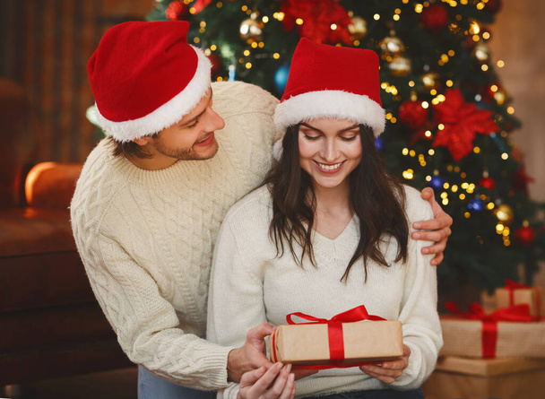 fröhlicher Mann mit Weihnachtsmütze begrüßt Frau mit Weihnachtsgeschenk - Foto, Bild