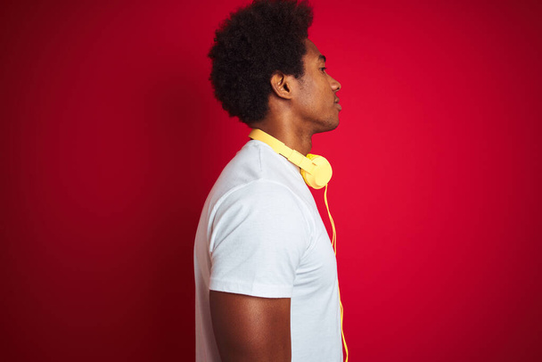Young african american man listening to music using headphones over isolated red background looking to side, relax profile pose with natural face with confident smile. - Valokuva, kuva