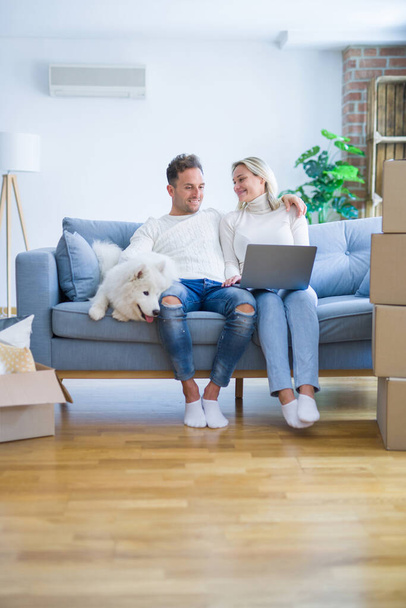 junges schönes Paar mit Hund sitzt mit Laptop auf dem Sofa im neuen Zuhause um Kartons herum - Foto, Bild