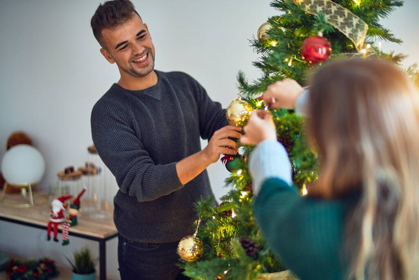 Молодая красивая пара улыбается счастливой и уверенной. Установка и украшение новогодней елки дома
 - Фото, изображение