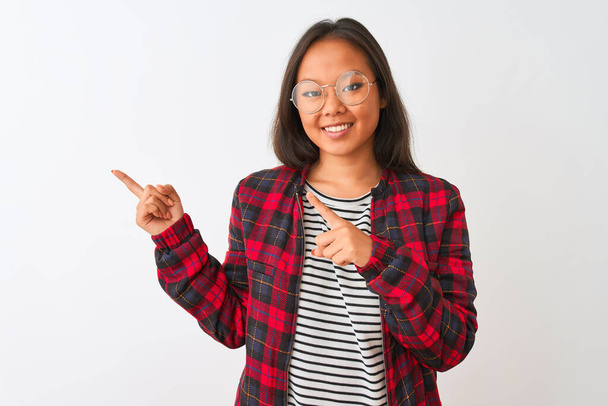 Jonge Chinese vrouw draagt t-shirt jas en bril over geïsoleerde witte achtergrond glimlachend en kijkend naar de camera wijzend met twee handen en vingers naar de zijkant. - Foto, afbeelding