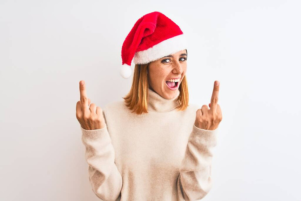 孤立した背景の上にクリスマスの帽子をかぶって美しい赤毛の女性あなたの悪い表情、挑発、失礼な態度をファックする中指を示しています。興奮して悲鳴 - 写真・画像