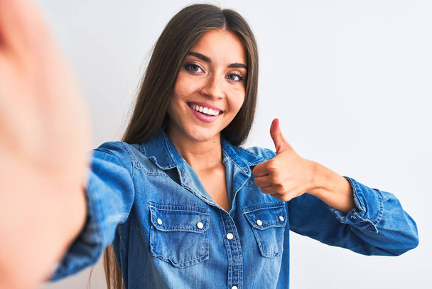 Beautiful woman wearing denim shirt make selfie by camera over isolated white background doing happy thumbs up gesture with hand. Approving expression looking at the camera showing success. - 写真・画像