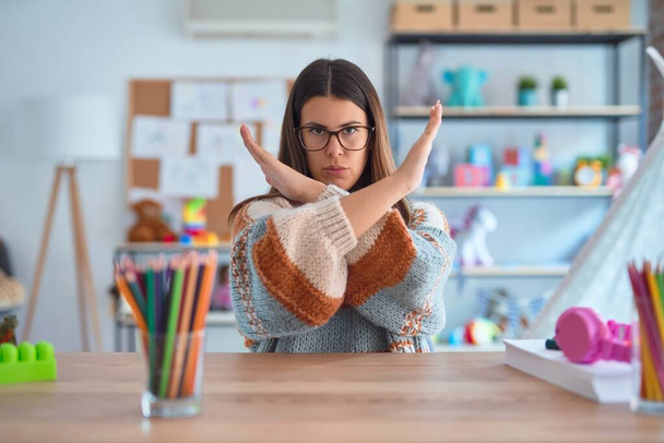 Young beautiful teacher woman wearing sweater and glasses sitting on desk at kindergarten Rejection expression crossing arms doing negative sign, angry face - Photo, Image