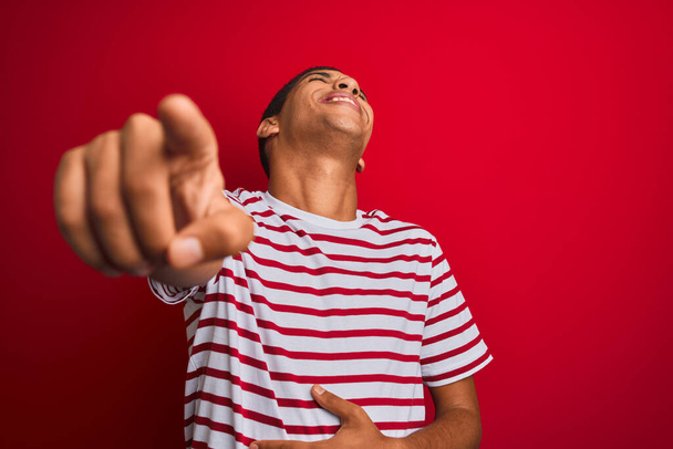 Νεαρός όμορφος Άραβας με ριγέ μπλουζάκι πάνω από απομονωμένο κόκκινο φόντο γελώντας μαζί σου, δείχνοντας την κάμερα με το χέρι πάνω από το σώμα, έκφραση ντροπής - Φωτογραφία, εικόνα