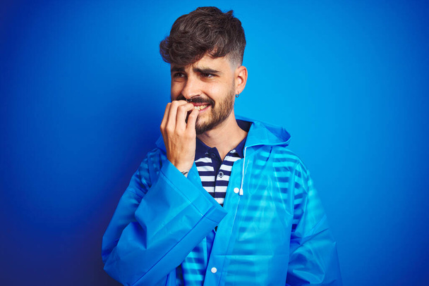 Молодой красивый мужчина в дождевой куртке, стоящий на изолированном синем фоне, выглядящий напряженным и нервным с руками на губах, грызущими ногти. Проблема тревоги
. - Фото, изображение
