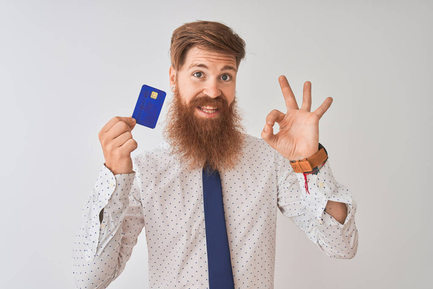 Jeune rousse irlandaise homme d'affaires tenant la carte de crédit debout sur fond blanc isolé faisant signe ok avec les doigts, excellent symbole
 - Photo, image