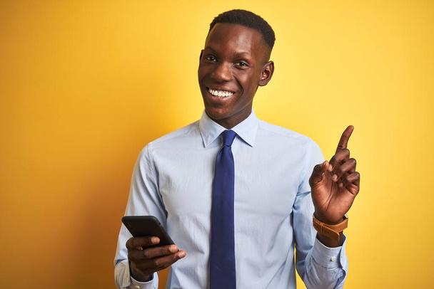 Afrika kökenli Amerikalı iş adamı, izole edilmiş sarı arka planda duran akıllı telefonu kullanıyor. Elini ve parmağını kenarı işaret ederek çok mutlu. - Fotoğraf, Görsel