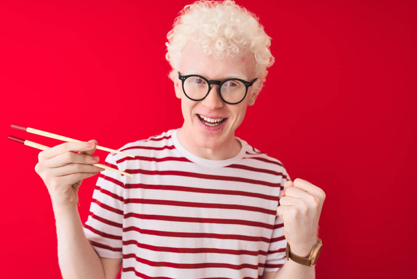 Young albino blond man holding chopsticks standing over isolated white background screaming proud and celebrating victory and success very excited, cheering emotion - 写真・画像